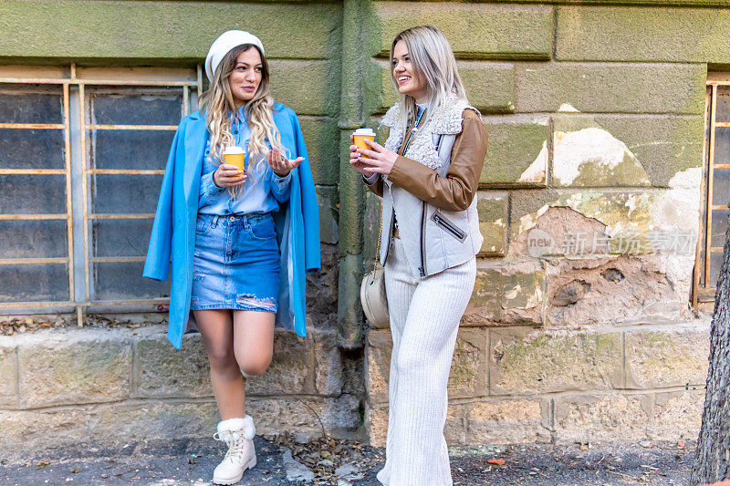 两个年轻时尚的女人，姐妹走在街上，拿着咖啡杯。微笑的姐妹在户外散步