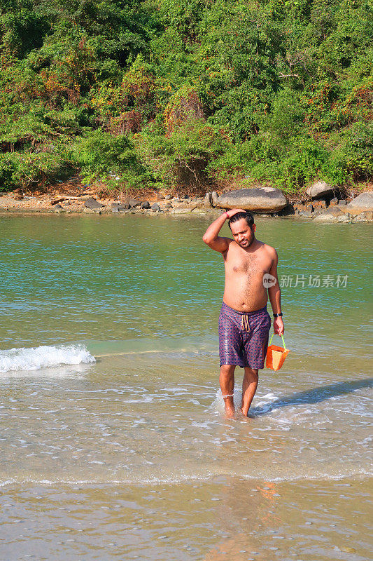 一名印度男子在海滩度假，背着塑料桶，从印度洋返回海滩，海浪翻滚到沙滩上，印度果阿巴洛伦海滩