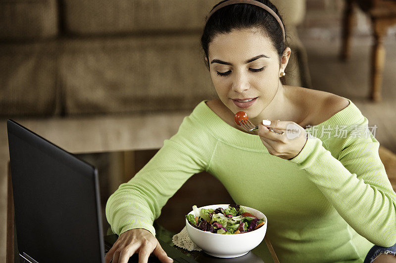 特写的西班牙裔可爱的年轻女子吃素食沙拉在午餐时使用笔记本电脑