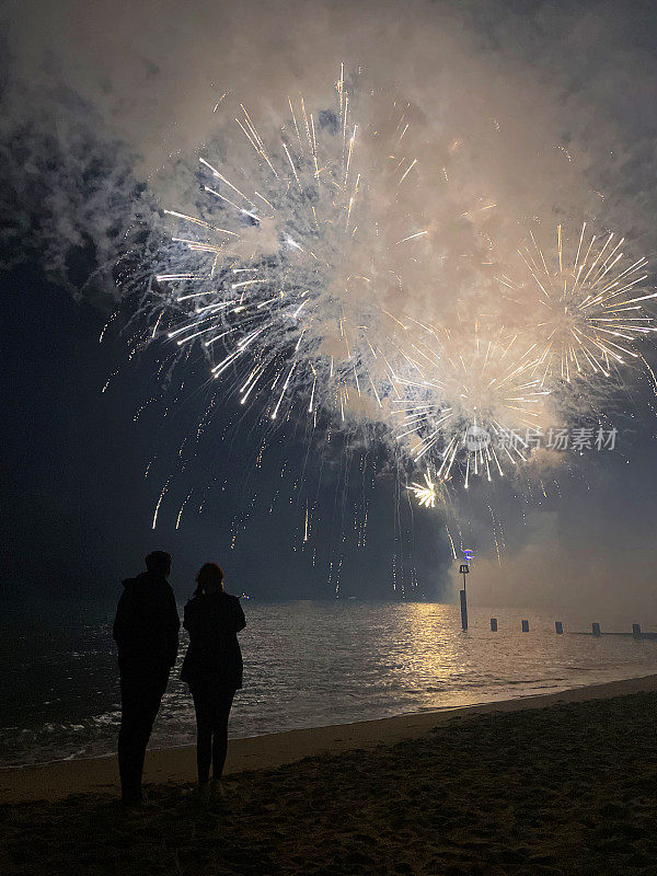 年轻夫妇的剪影在海滩上站在水边观看烟花表演，烟雾和火花在夜空中