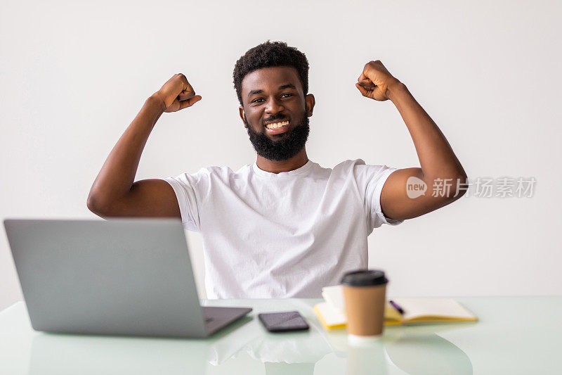 快乐的非洲商人在西装看着笔记本电脑兴奋的好消息在线。黑人胜者坐在办公桌前达成目标，举起双手庆祝事业成功，赢得胜利