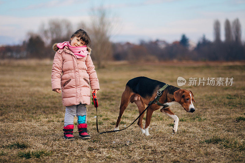 一个美丽的小女孩站在草地上，用皮带牵着她的狗。他们喜欢散步。