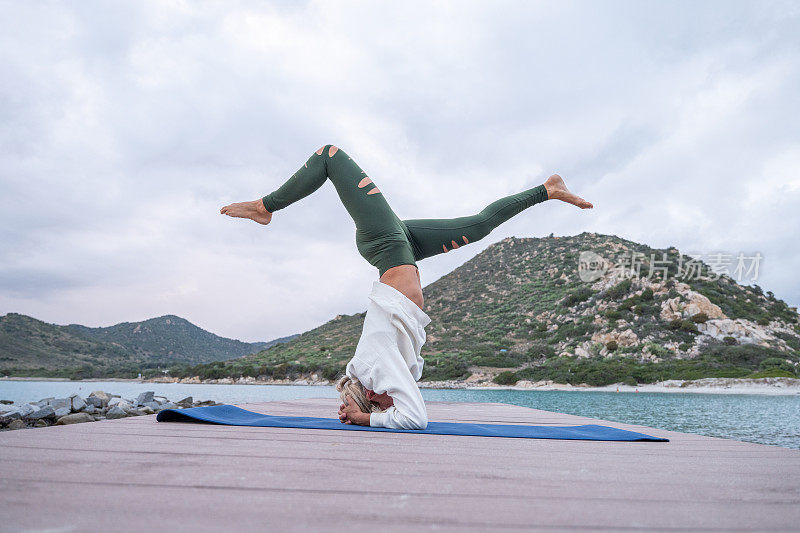 灵活的年轻女子练习瑜伽倒置姿势在一个码头在早上。倒立，倒立，撒丁岛的海景