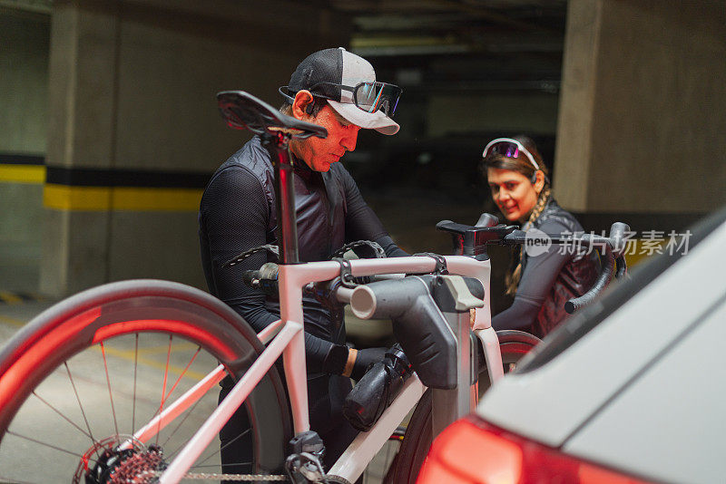 穿着自行车装备的拉丁裔异性恋夫妇穿过他们大楼的公共空间，把自行车拿到车上，然后去他们要骑的地方