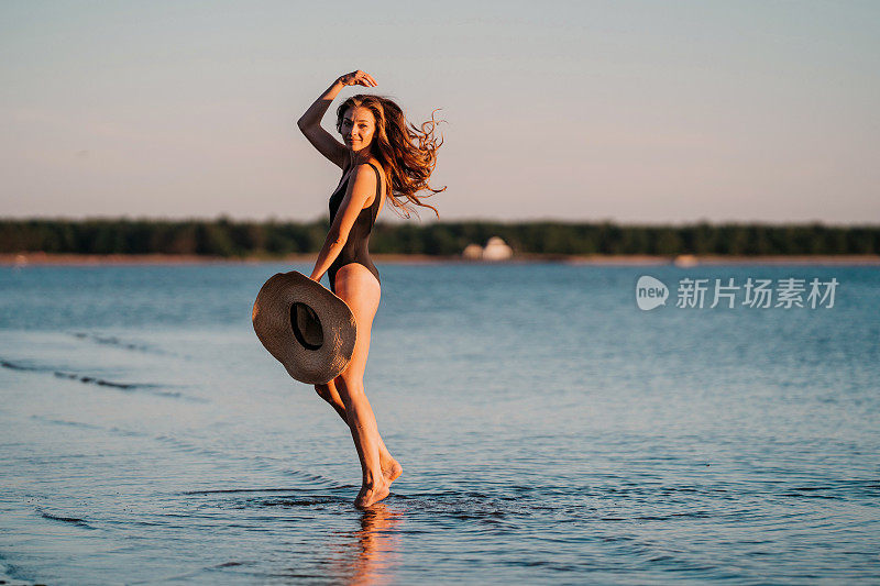 华丽的年轻适合意大利女子在泳装走在海滩上手持草帽，日落时分。成功的奢华女性度假时在岛上放松，享受海风。旅游、休闲活动