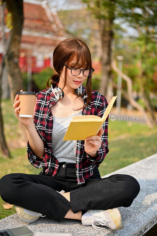 美丽的亚洲女大学生在公园里喝着咖啡，专注地看书。
