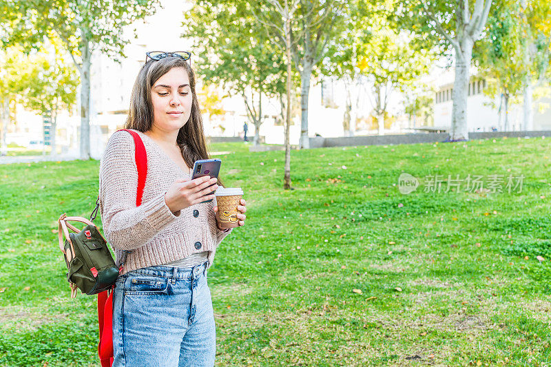 侧视图一个微笑的女人用智能手机咖啡站在公园里