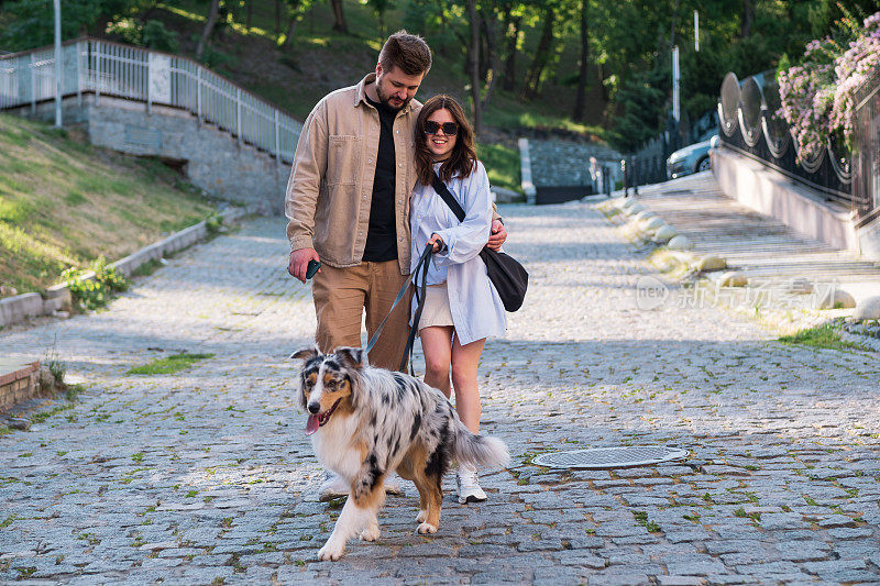 一男一女带着漂亮的澳大利亚牧羊犬在市区散步。幸福的夫妇和他们的宠物在城市里，带着狗去市中心