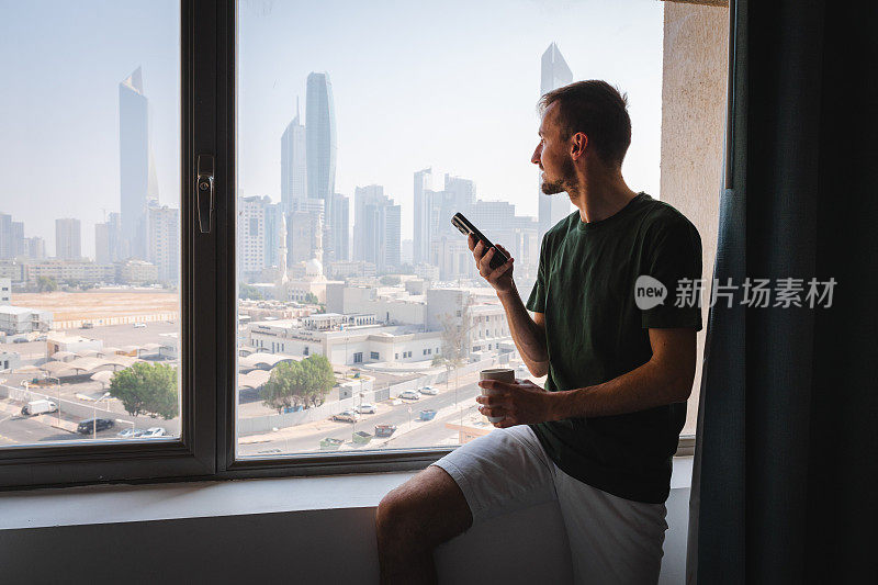 一名年轻的白人男子在酒店房间边看窗外边玩手机