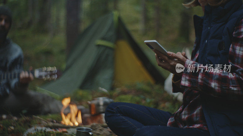 度假期间，女人在篝火边玩智能手机，而男人在森林里弹吉他