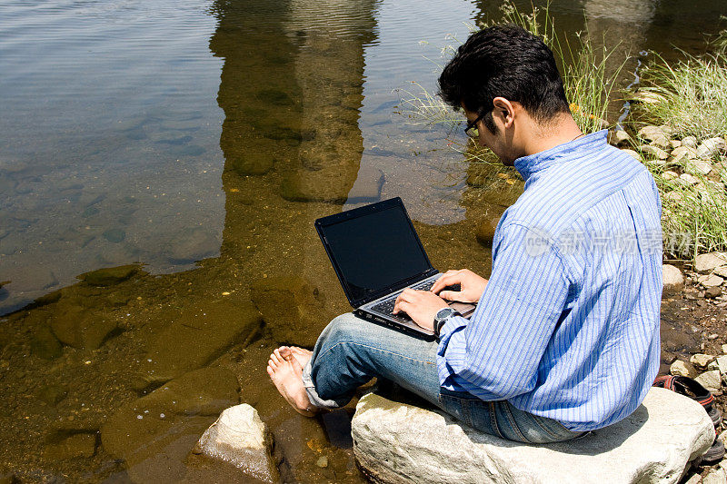印度青年使用笔记本电脑工作在户外水域附近