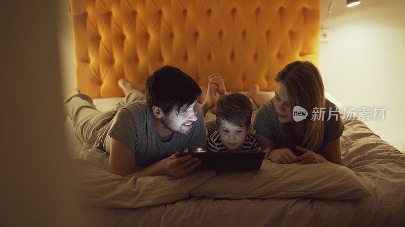 一个幸福的家庭，小儿子躺在家里的床上，睡觉前用平板电脑看卡通电影