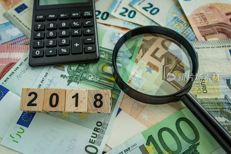 财税概念为欧元钞票堆上的放大镜，计算器和木块上的2018号