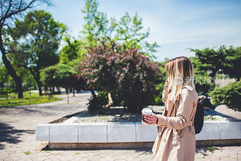 一个女人在城市公园里走着手里拿着咖啡一边讲着手机。