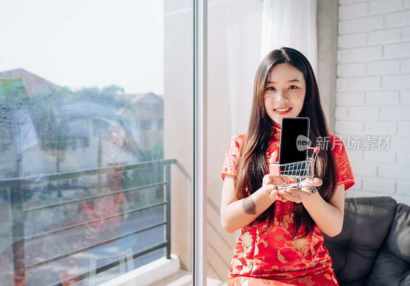 购物亚洲女人中国服装，旗袍，旗袍春节和抱购物车，智能手机坐在靠近窗户在干净的白色房间，拷贝空间。