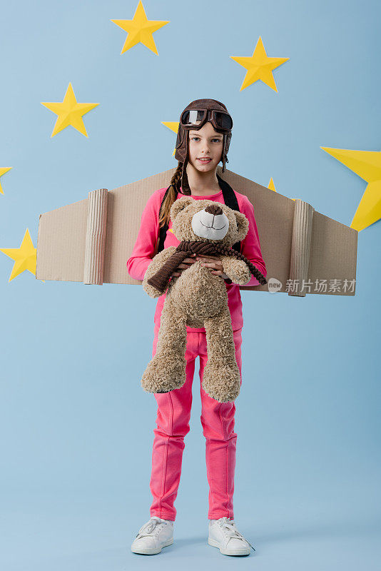 好奇的孩子用纸板翅膀抱着蓝色星空背景下的泰迪熊