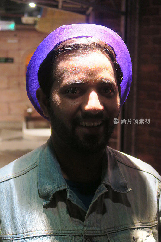 英俊的年轻印度男子在20岁出头微笑着，戴着一顶紫色的帽子，修剪整齐的胡子，穿着淡蓝色牛仔夹克，华丽的印度男子站在迪斯科灯光下给阴影效果，摆出性感的姿势看着镜头。