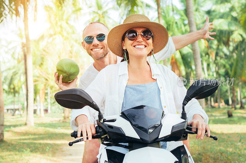 快乐的微笑夫妇旅行者骑摩托车踏板车在棕榈树下。一个笑着拿着椰子的男人。热带度假概念形象