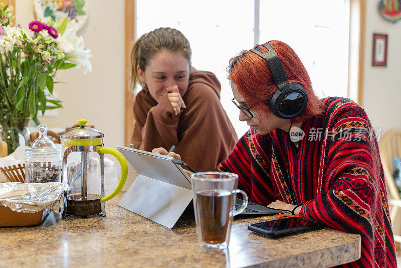 当你在家学习的时候，你是一个更新的人。一个十几岁的女孩戴着耳机在远处上课，在厨房柜台边用笔记本电脑工作，她的姐姐在一旁看着，可能会分散她的注意力。