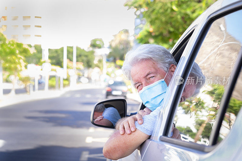 有魅力的微笑白发老人坐在车里戴着外科口罩，他的胳膊靠在车窗上，看着镜头