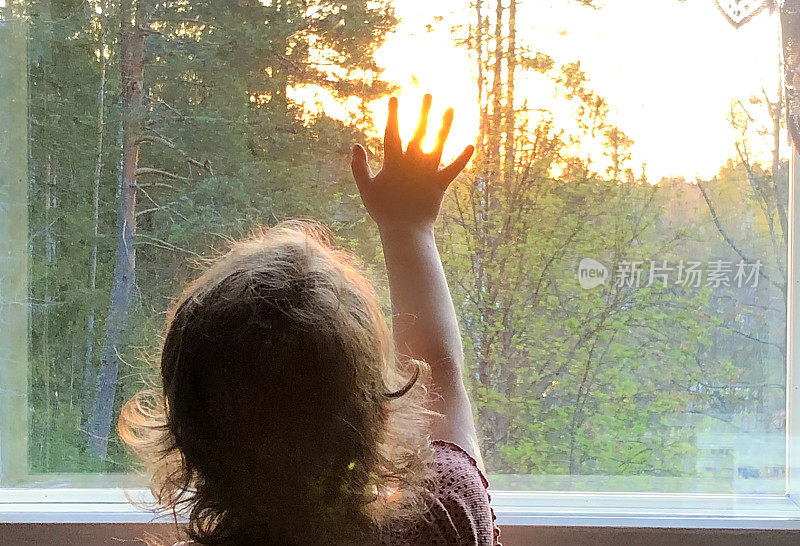 一个5岁的小女孩站在窗前看日落。从后面可以看到这个孩子。孩子用他的手抓住了太阳和梦想。
