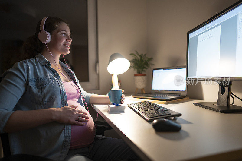 怀孕的女商人在她的家庭办公室工作到很晚，戴着耳机听音乐