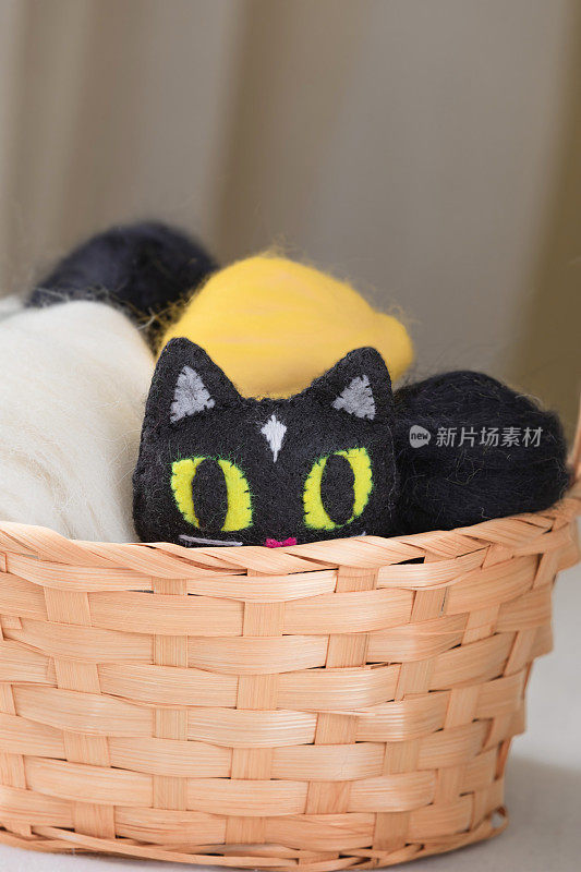 黄色眼睛的毛毡玩具黑猫藏在柳条篮子里，米黄色背景上有毛球。手工制作的创意软玩具小猫。