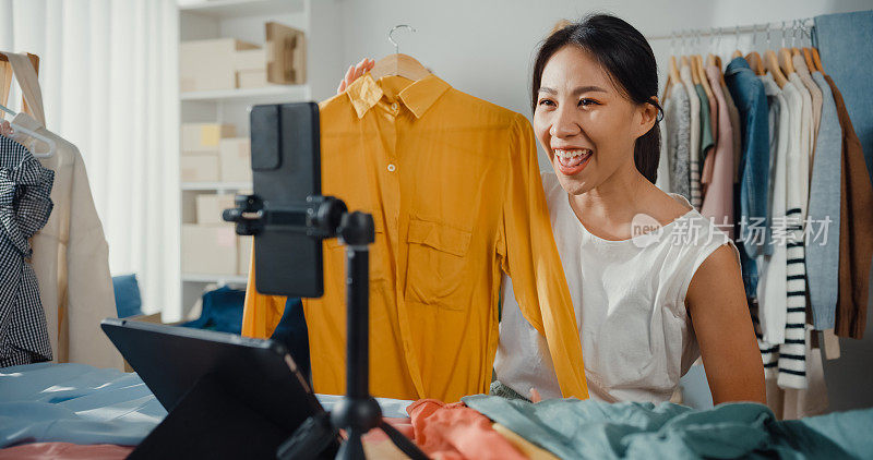 亚洲女商人直播电子商务在家里卖衣服，美丽的女孩使用智能手机和平板电脑录制视频。