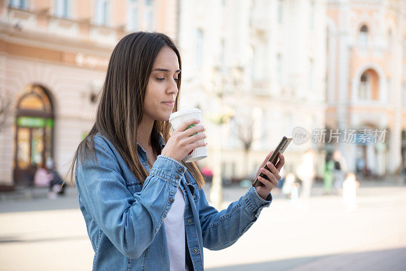 快乐的年轻女子走在城市的街道上，用她的智能手机