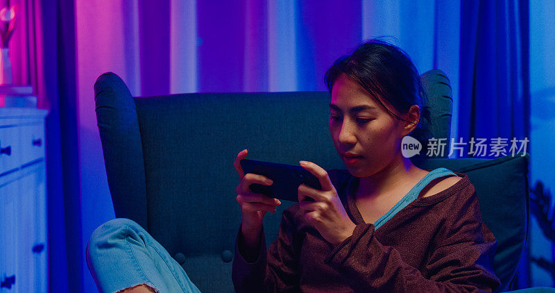 咄咄逼人的亚洲女人与休闲衬衫在沙发椅子上戴耳机拿智能手机感到心烦生气的失败者竞争玩网络游戏坐在客厅的家在晚上。