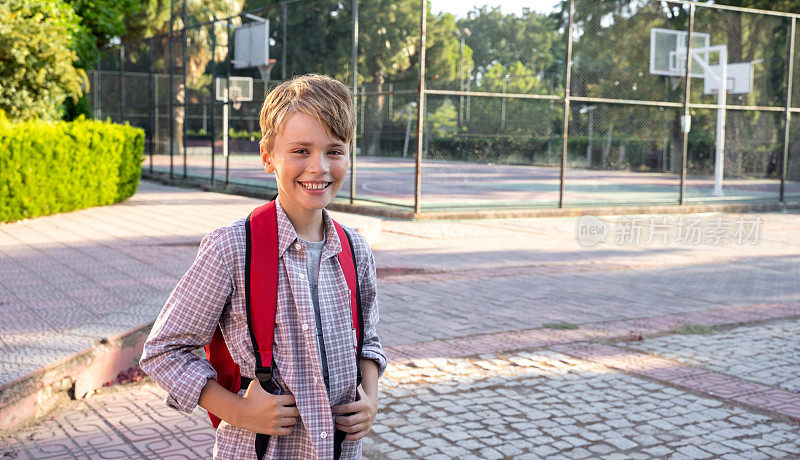 中学男生背着书包对着镜头微笑，体育课结束后在操场上打排球。
