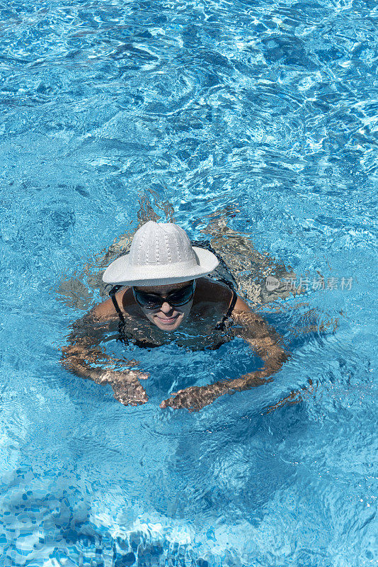 戴着白色帽子和太阳镜的成年女子正在蓝色的水中游泳。