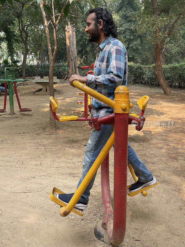 印度男子的形象在牛仔裤和衬衫工作在户外健身房使用空气行者健身运动设备，锻炼户外露天公园，侧面视图