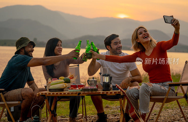 美丽的高加索女人和她的朋友在露营时用手机自拍，在日落的湖边一起享受，他们看起来很高兴参加户外派对。