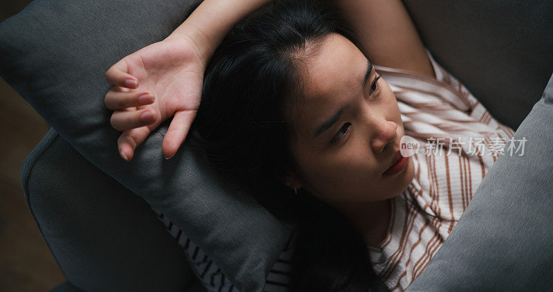 忧郁沮丧的年轻亚洲女子躺在家里的沙发上，看着天花板思考着