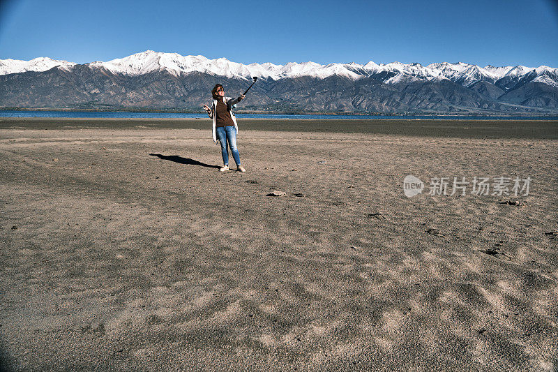一名女游客走在山间湖岸，用运动相机拍照