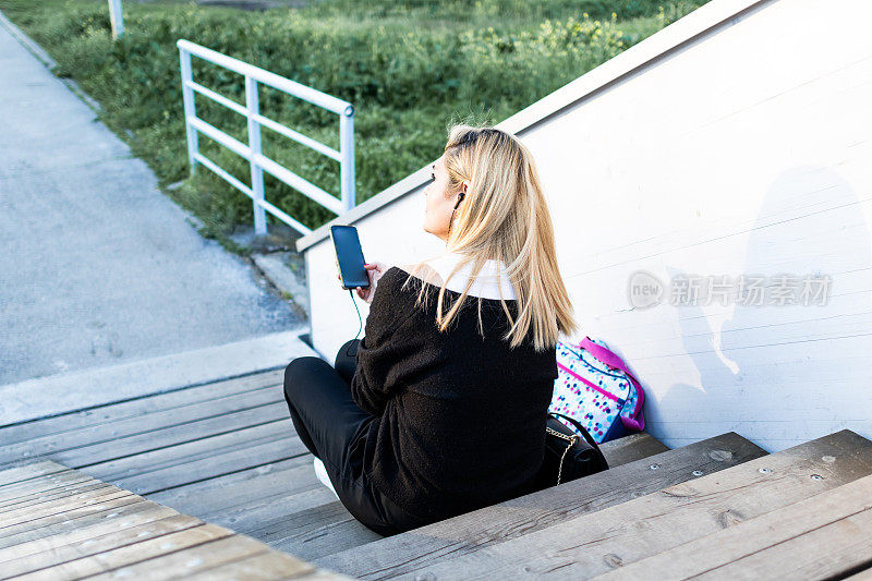 一个美丽的女商人的肖像，下班后在楼梯上享受户外，听音乐。商务人士带着电子设备上下班。