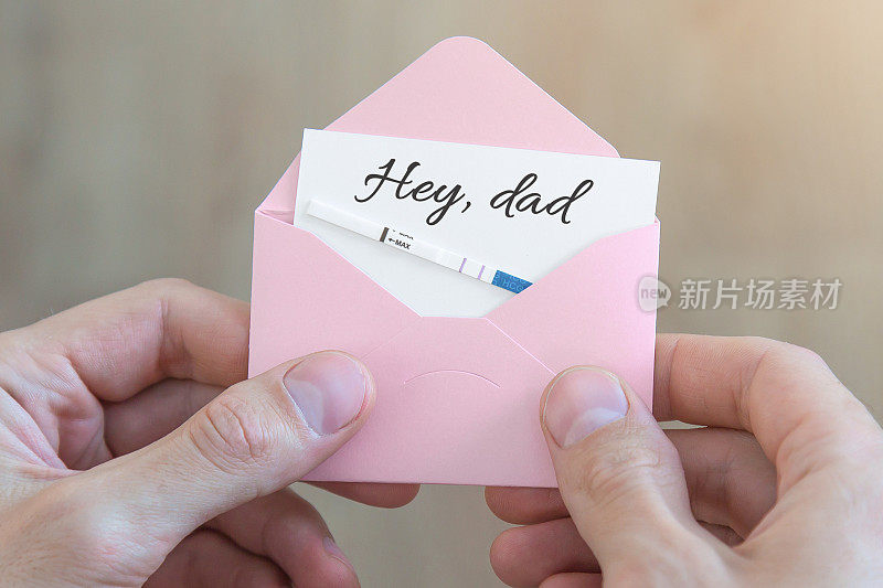 男性手里拿着一个粉红色的信封，里面有一张明信片，上面写着“嘿，爸爸，怀孕测试呈阳性，怀孕的概念。