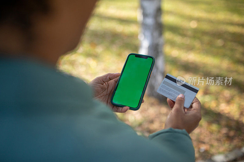 从肩膀上看，一个女人拿着绿屏手机，另一只手拿着信用卡