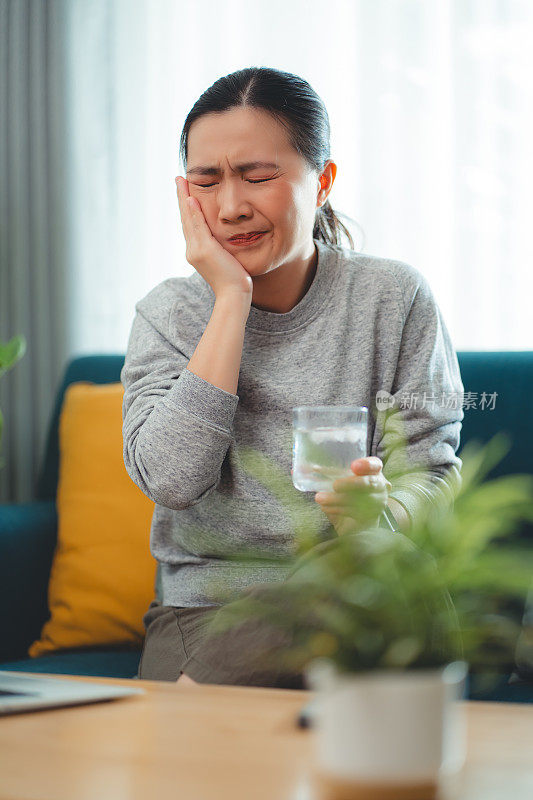 亚洲女性坐在客厅的沙发上，喝了凉水，摸了摸脸颊，就会感到牙齿敏感。