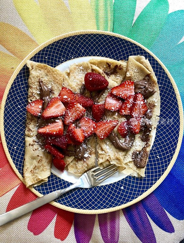 想象一下自制的薄饼早餐，配上草莓片，融化的榛子巧克力酱和干椰子，用叉子放在蓝色和金色的餐盘里