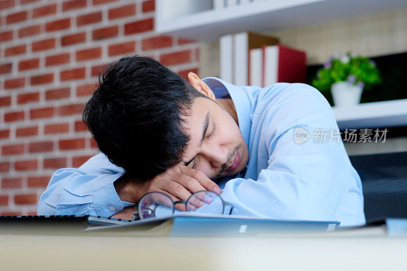 疲惫的亚洲人在办公桌前睡觉。戴着眼镜的年轻商人过度劳累而睡着了，创造性的休闲男人在他的工作场所睡觉