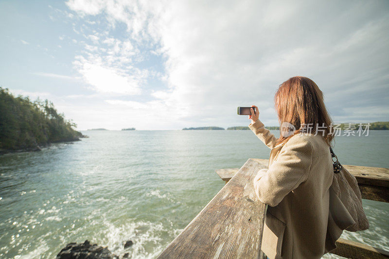 年轻女子在海边的铁轨上用手机拍摄风景