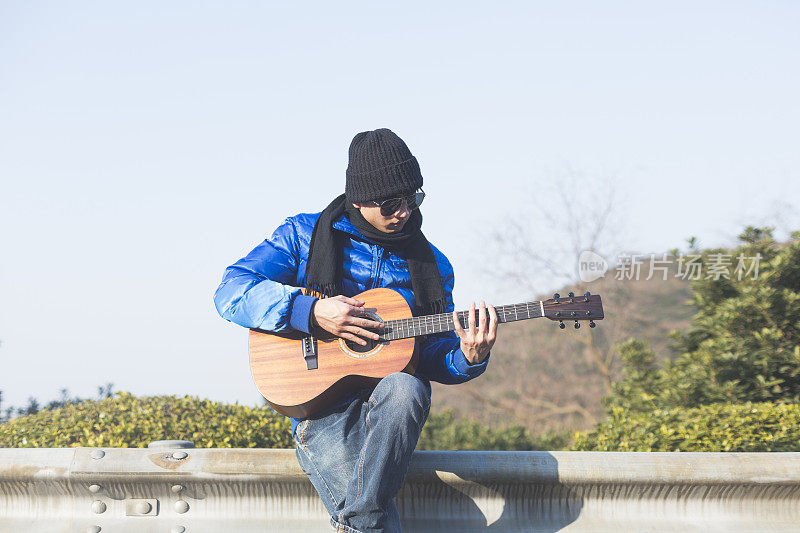 年轻的亚洲男子坐在公路栏杆上弹吉他