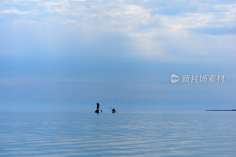 站立式划桨板和皮划艇的剪影在平坦温暖安静的海面上划桨。两个水上游客站着划桨板(SUP)和黄色皮划艇划桨在一个平坦温暖安静的海上