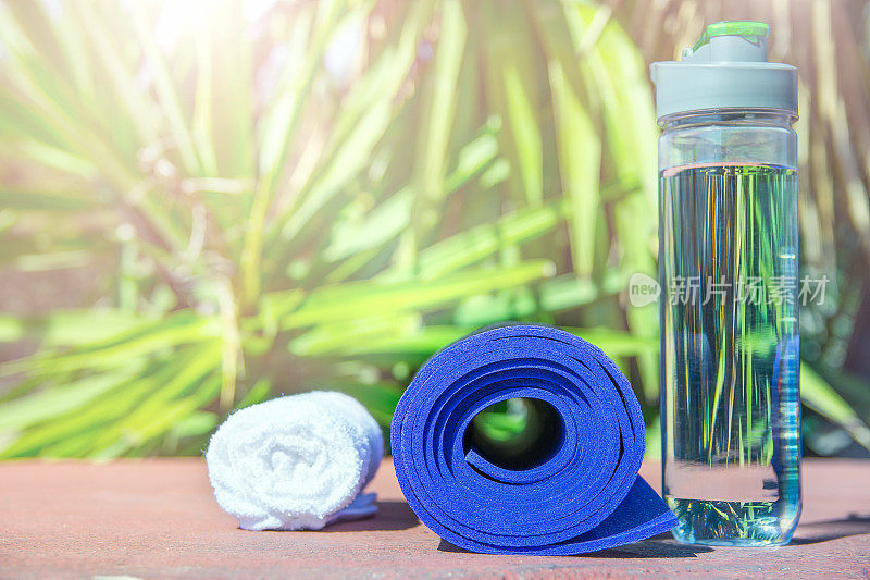 蓝色卷瑜伽垫瓶与水白色毛巾在绿色棕榈树自然背景。明亮的正午的阳光。放松夏季冥想健身幸福概念。本空间