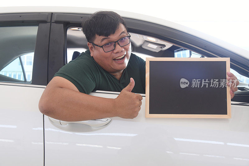 一名亚裔男子坐在车里，手里拿着一张黑色空白海报，竖起大拇指。