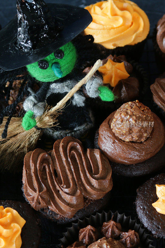 这是万圣节女巫和扫帚的特写，上面是自制的巧克力纸杯蛋糕，上面是棕色和橙色的管道糖霜，黑色的纸蛋糕盒在黑色的背景上，俯瞰