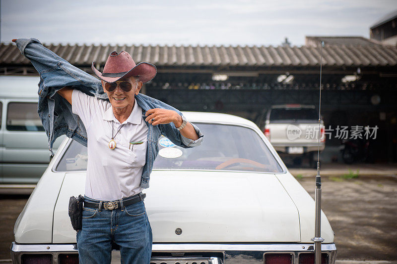 年长的亚洲男人在牛仔夹克摆姿势与古董车