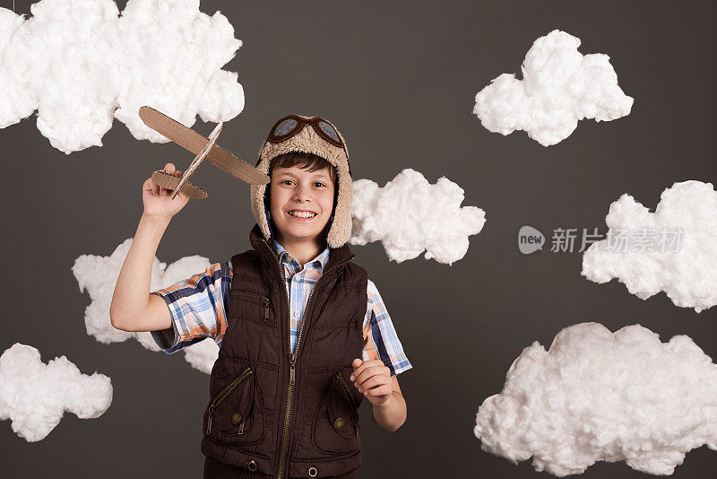 一个男孩在玩纸板飞机，梦想成为一名飞行员，穿着复古风格的夹克，戴着头盔和眼镜，棉花云，灰色的背景，色调为棕色